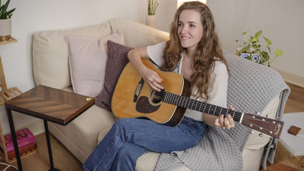 A woman with long wavy hair sits on a couch playing an acoustic guitar. She is smiling and looking ahead. The cozy room, perfect for a guitar lesson, features a couch adorned with a knitted gray blanket and colorful cushions. A small table with a book and some plants adds warmth to the scene.